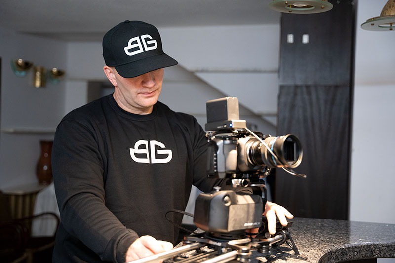 Kamerzysta w czarnej bluzie i czapce z logo obsługuje profesjonalny sprzęt filmowy na statywie szynowym w jasnym wnętrzu. Gotowy do nagrywania.