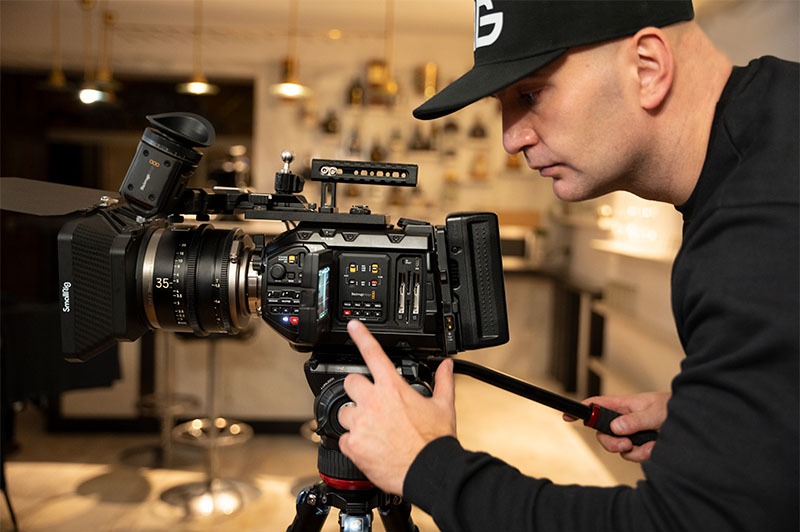 Operator z czapką reguluje ustawienia profesjonalnej kamery Blackmagic na statywie, z obiektywem 35mm, w jasnym wnętrzu. Skupienie na ekranie kamery.