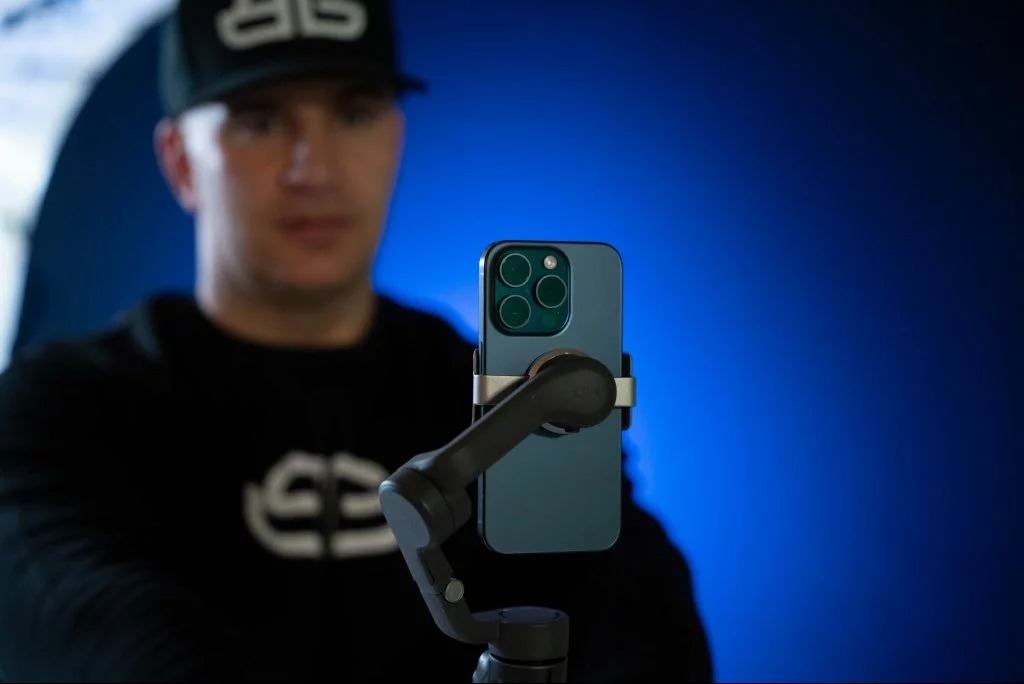 Mężczyzna z telefonem na stabilizatorze, nagrywa wideo na tle niebieskiego ekranu. Ujęcie z bliska, widoczny sprzęt do filmowania.
