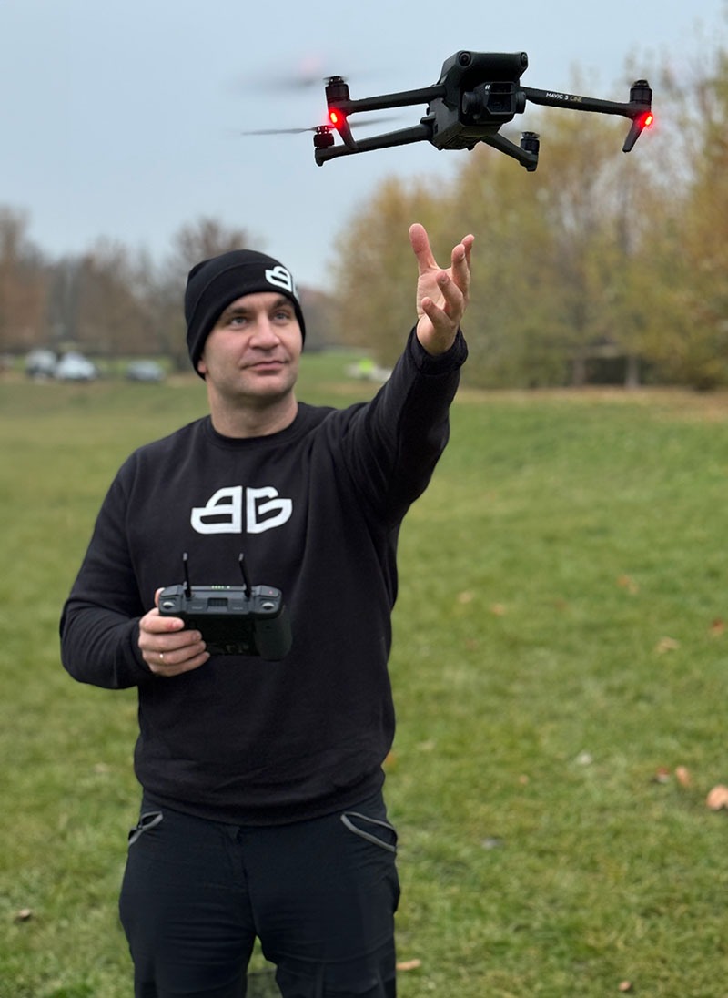 Mężczyzna w czarnej bluzie i czapce steruje dronem Mavic 3 Cine na tle jesiennego krajobrazu. Dron z czerwonymi światłami w locie.