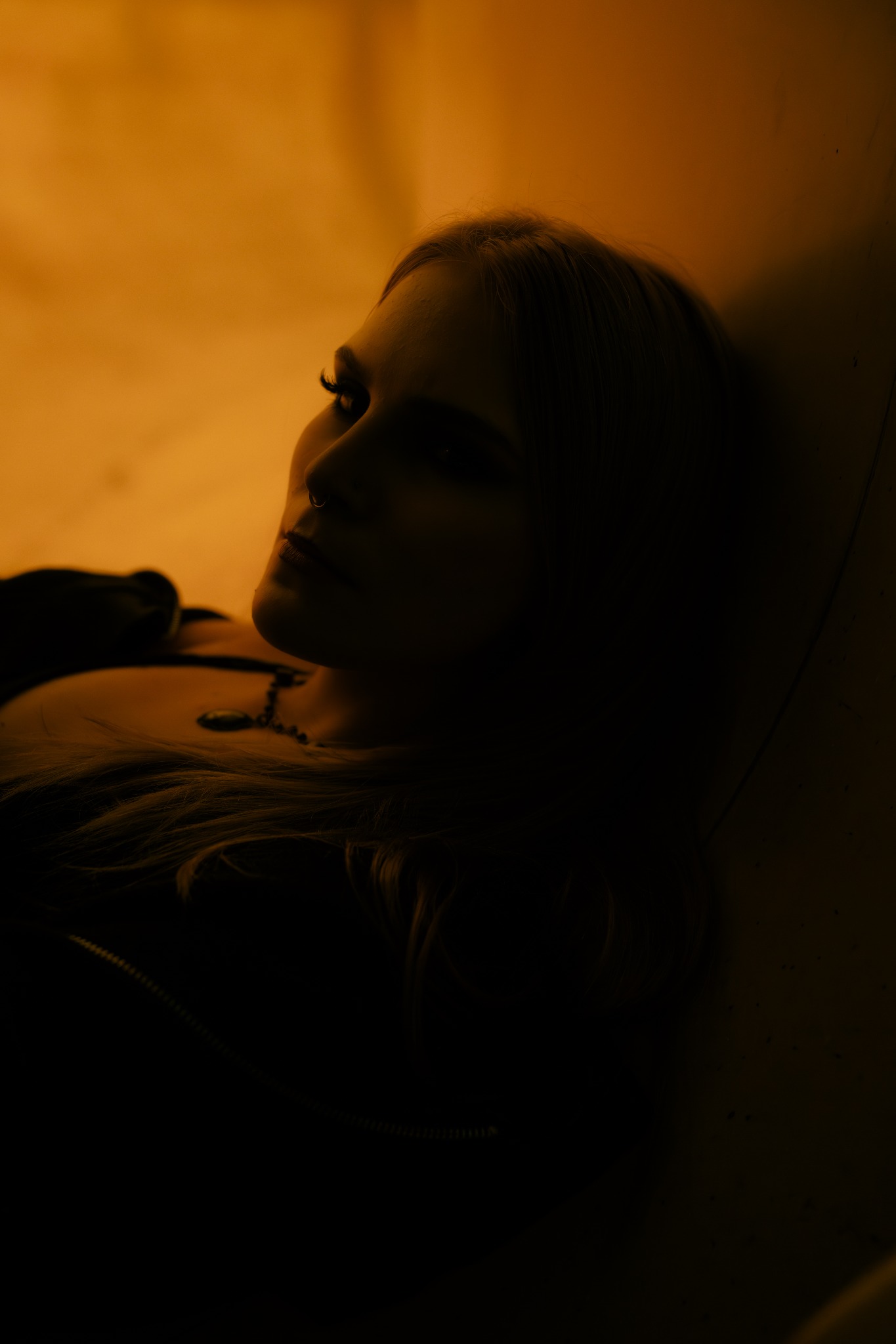 Portret kobiety w półmroku, z delikatnym oświetleniem podkreślającym rysy twarzy i subtelny makijaż. Widoczny piercing nosa i naszyjnik. Klimatyczne, artystyczne ujęcie.