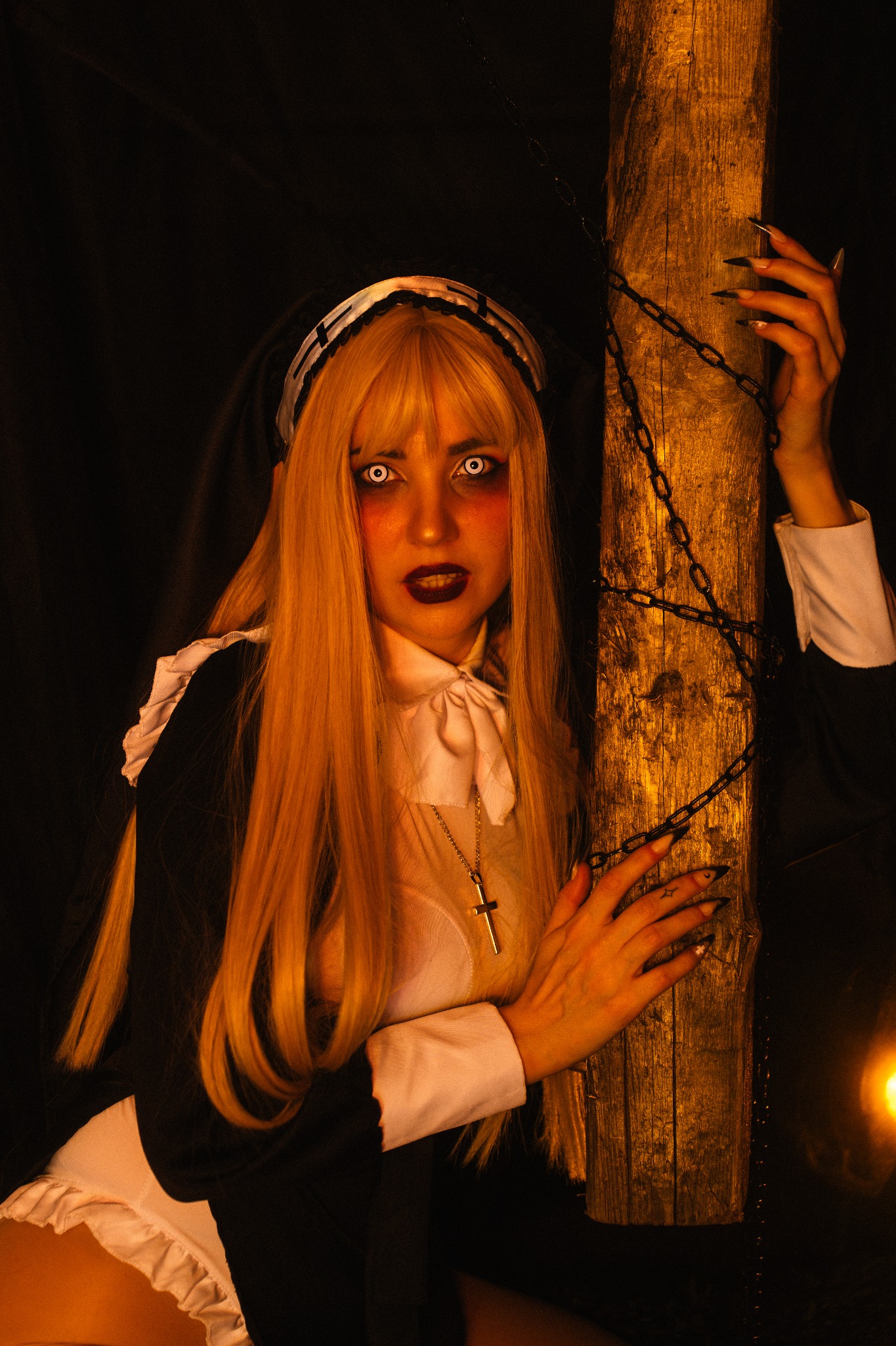 Portret kobiety w kostiumie zakonnicy z upiornym makijażem, opierającej się o drewniany słup opleciony łańcuchem, w mrocznym oświetleniu.
