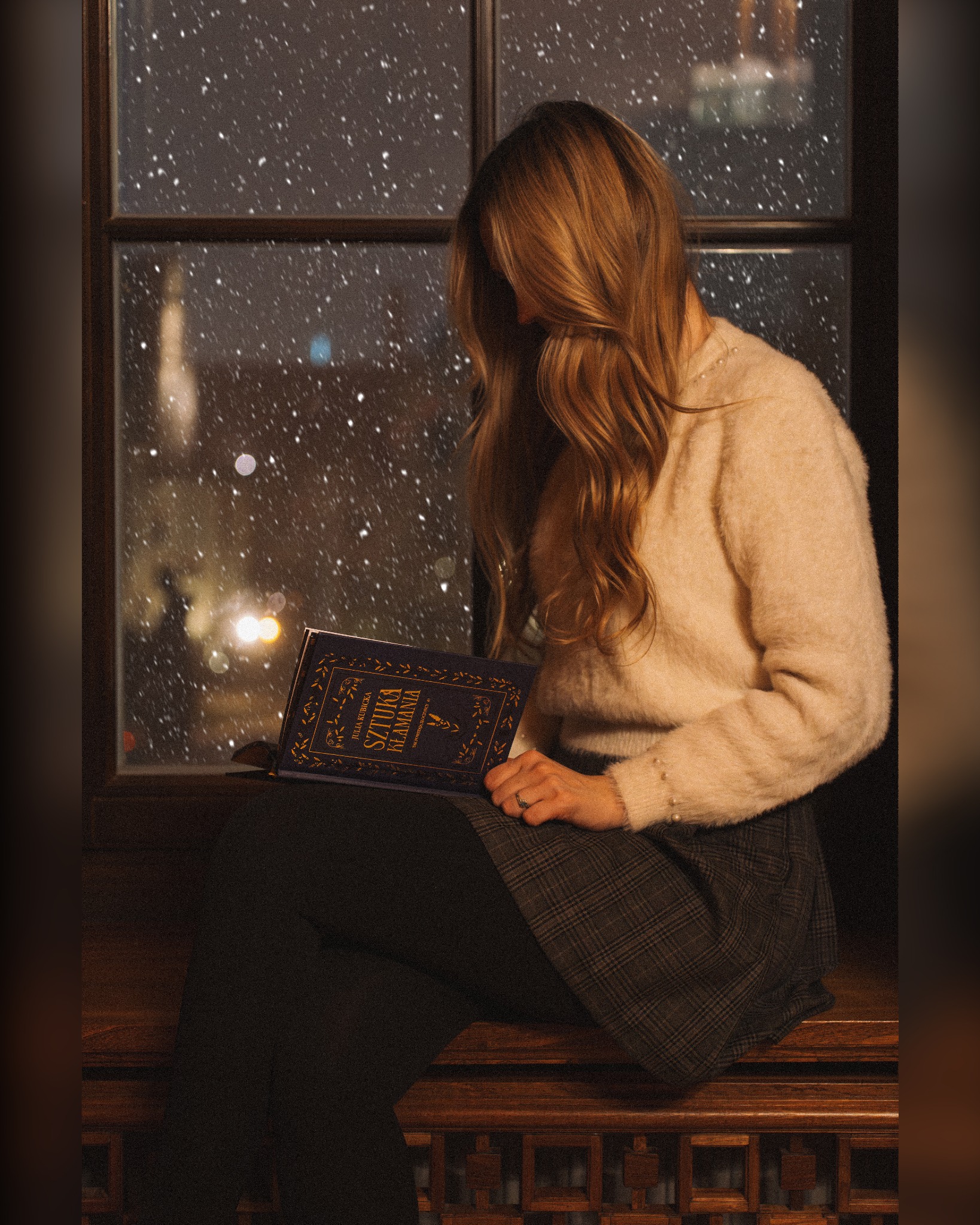 Kobieta z książką 'Sztuka kłamania' siedzi na parapecie w Poznaniu, obserwując padający śnieg za oknem. Klimatyczna sesja w zimowy wieczór.