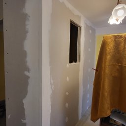 Usługi Remontowo- Budowlane Piotr Sądej - Ściana z płyt karton-gips z widocznymi miejscami na szpachlowanie, fragment okna. Zabezpieczenie mebli złotą tkaniną. Etap prac wykończeniowych w pomieszczeniu.