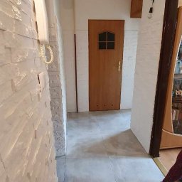 Usługi Remontowo- Budowlane Piotr Sądej - Wykończony korytarz z białą ścianą z imitacji kamienia, szarymi płytkami podłogowymi i drewnianymi drzwiami. Widoczny fragment szafki i instalacji elektrycznej.