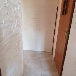 Usługi Remontowo- Budowlane Piotr Sądej - Korytarz po remoncie: biała cegła dekoracyjna na ścianach, szare płytki podłogowe, drewniane drzwi z szybką. Widok na odnowione wnętrze.