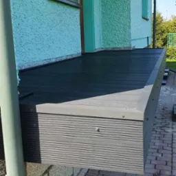 Nowoczesny balkon z kompozytowych desek w odcieniach szarości, z widoczną elewacją budynku w tle i fragmentem rynny. Kąt ujęcia z dołu podkreśla solidność konstrukcji.