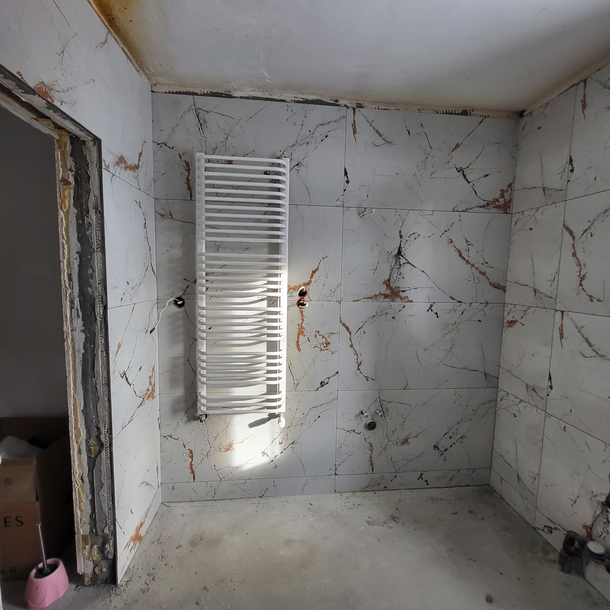Remont łazienki: Ułożone marmurowe płytki na ścianach, zamontowany biały grzejnik drabinkowy. Widoczne niedokończone wykończenia przy drzwiach.