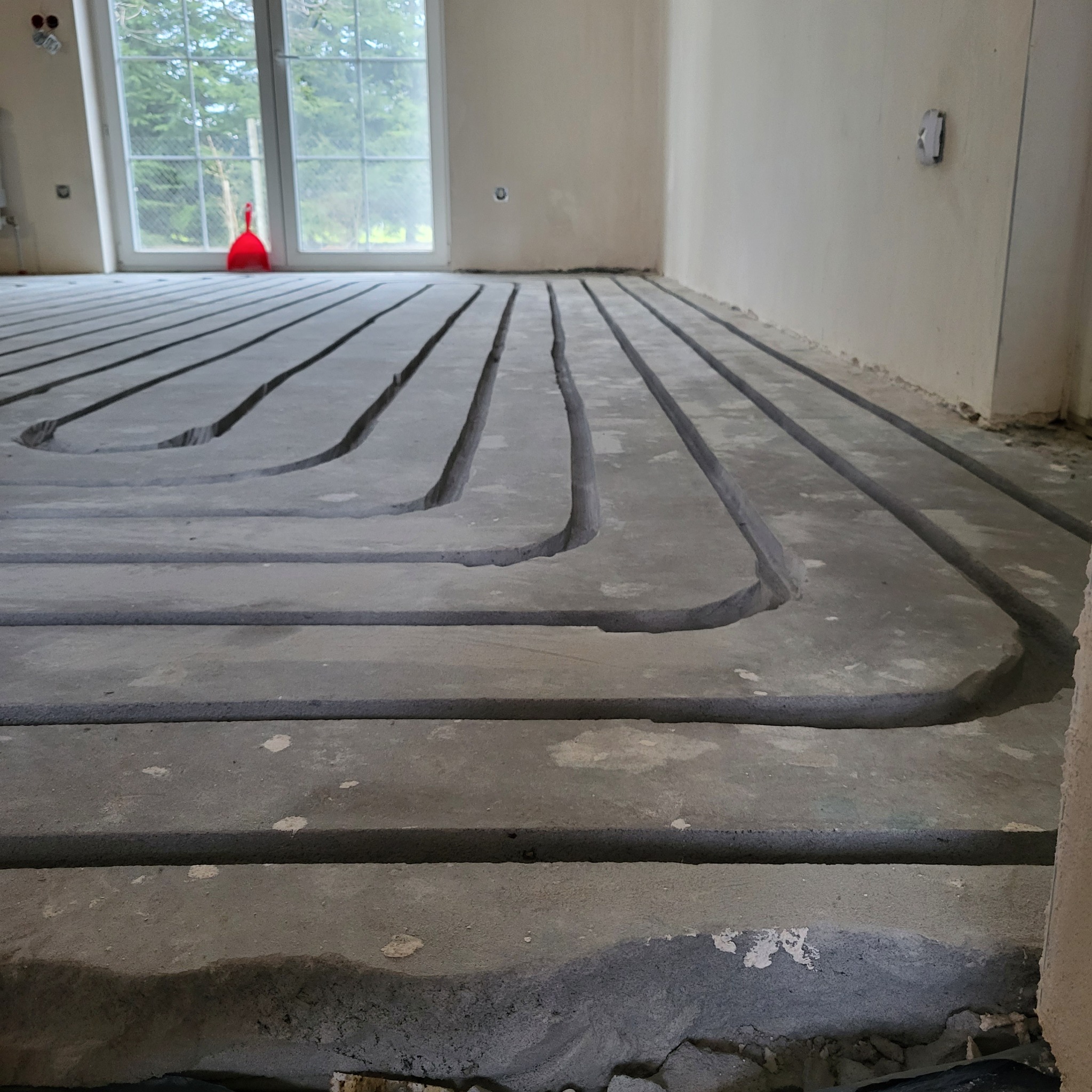 Przygotowanie do ogrzewania podłogowego: wyfrezowane rowki w podkładzie podłogowym, widoczne okno i resztki gruzu. Szare odcienie betonu.