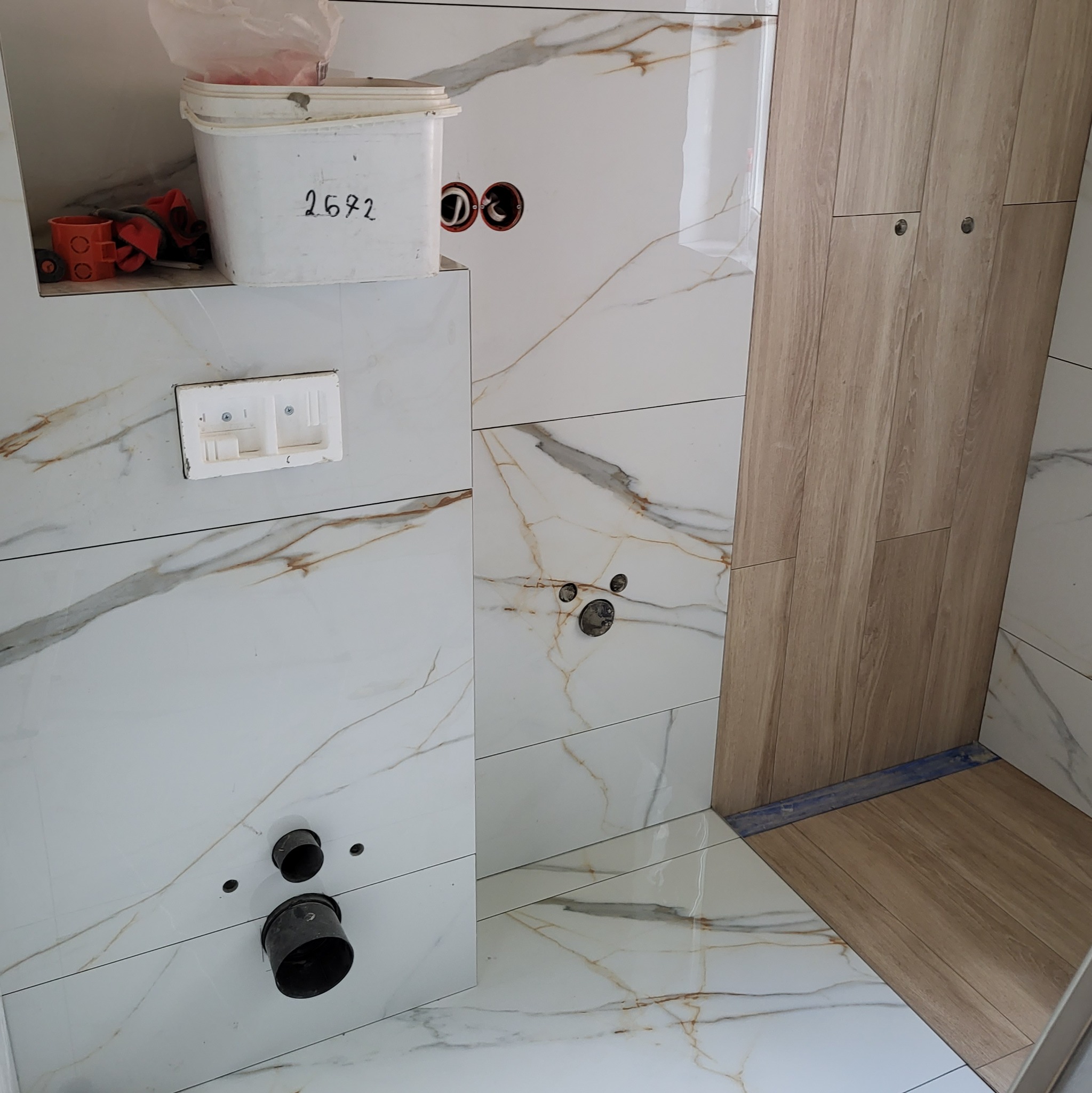 Białe płytki imitujące marmur z instalacją hydrauliczną i elektryczną w łazience w trakcie wykańczania, widoczny brodzik wyłożony płytkami drewnopodobnymi.