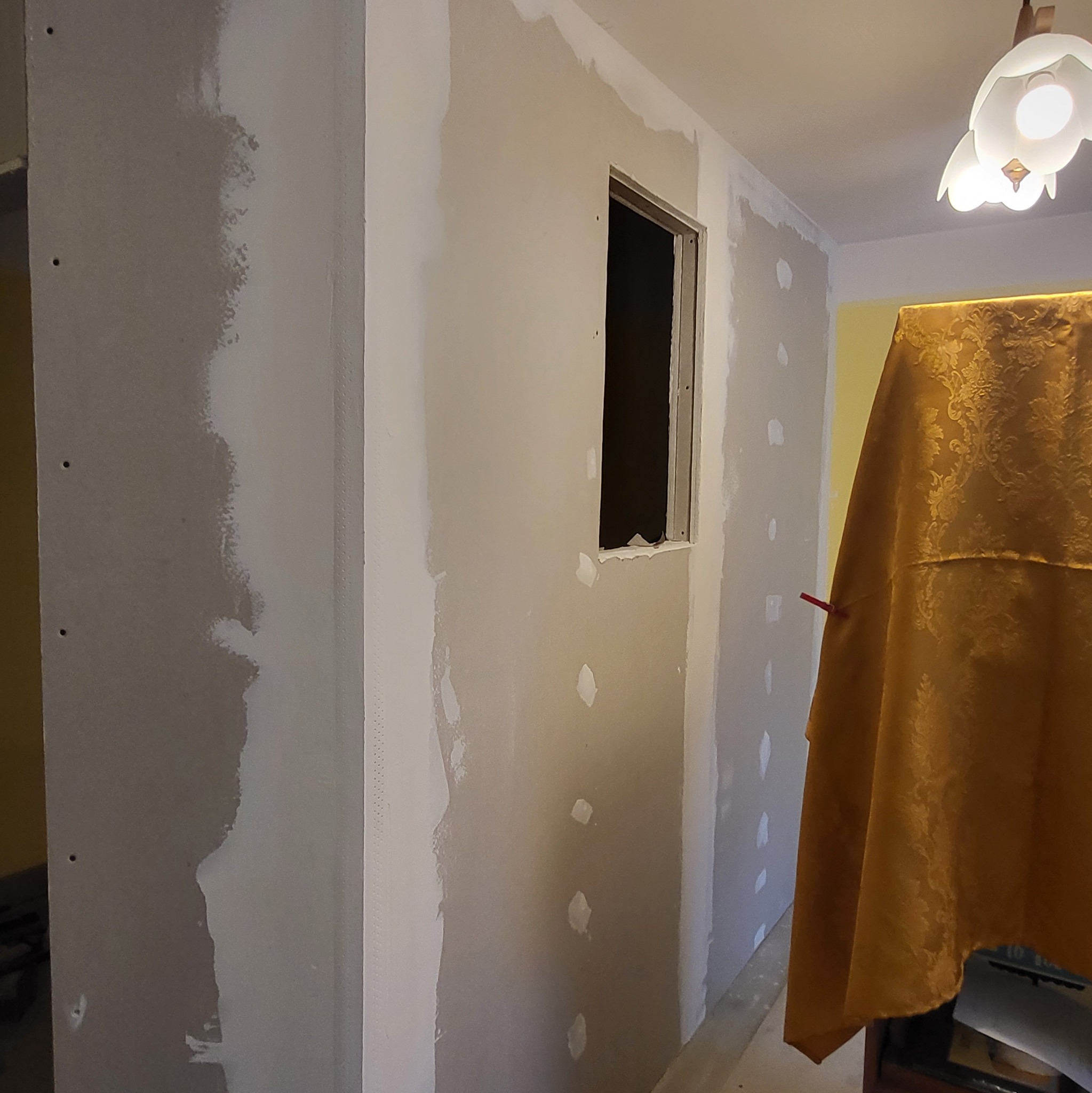 Ściana z płyt karton-gips z widocznymi miejscami na szpachlowanie, fragment okna. Zabezpieczenie mebli złotą tkaniną. Etap prac wykończeniowych w pomieszczeniu.