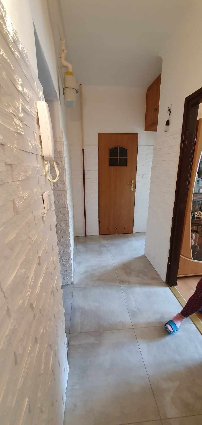 Wykończony korytarz z białą ścianą z imitacji kamienia, szarymi płytkami podłogowymi i drewnianymi drzwiami. Widoczny fragment szafki i instalacji elektrycznej.