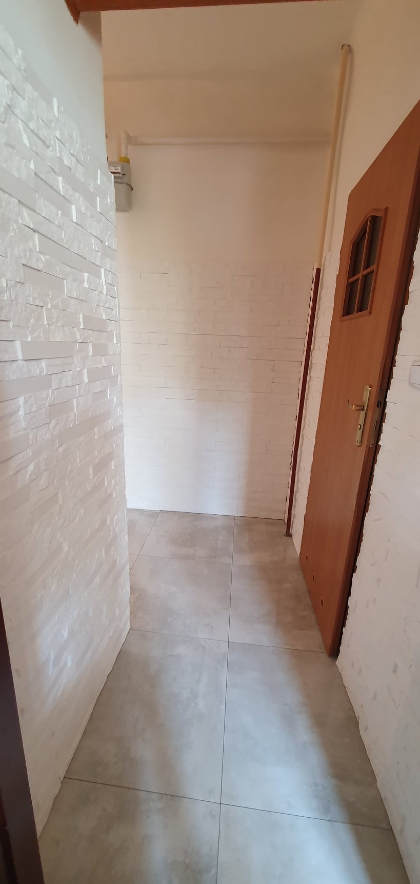 Korytarz po remoncie: biała cegła dekoracyjna na ścianach, szare płytki podłogowe, drewniane drzwi z szybką. Widok na odnowione wnętrze.