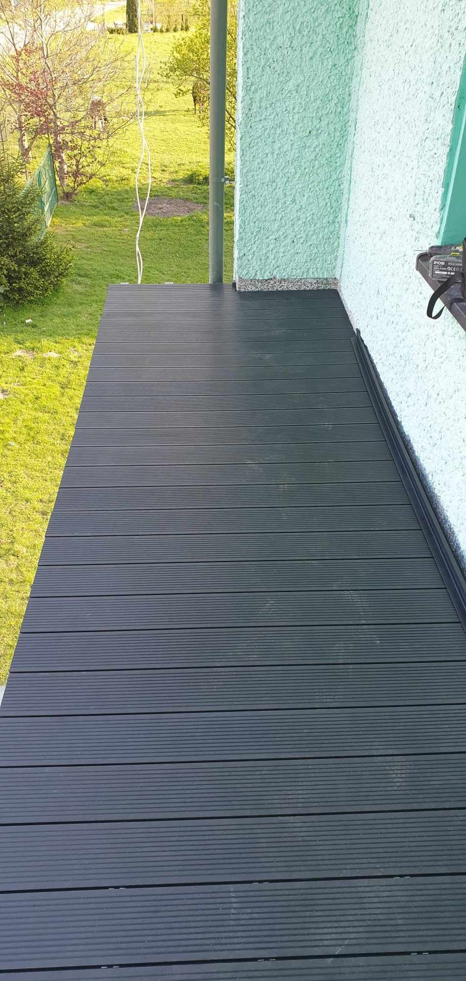 Nowoczesny balkon z ciemną, ryflowaną deską kompozytową, widok z góry na tle zieleni. Ściana budynku w jasnym odcieniu zieleni.