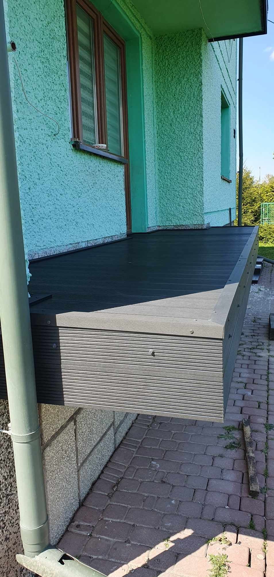 Nowoczesny balkon z kompozytowych desek w odcieniach szarości, z widoczną elewacją budynku w tle i fragmentem rynny. Kąt ujęcia z dołu podkreśla solidność konstrukcji.