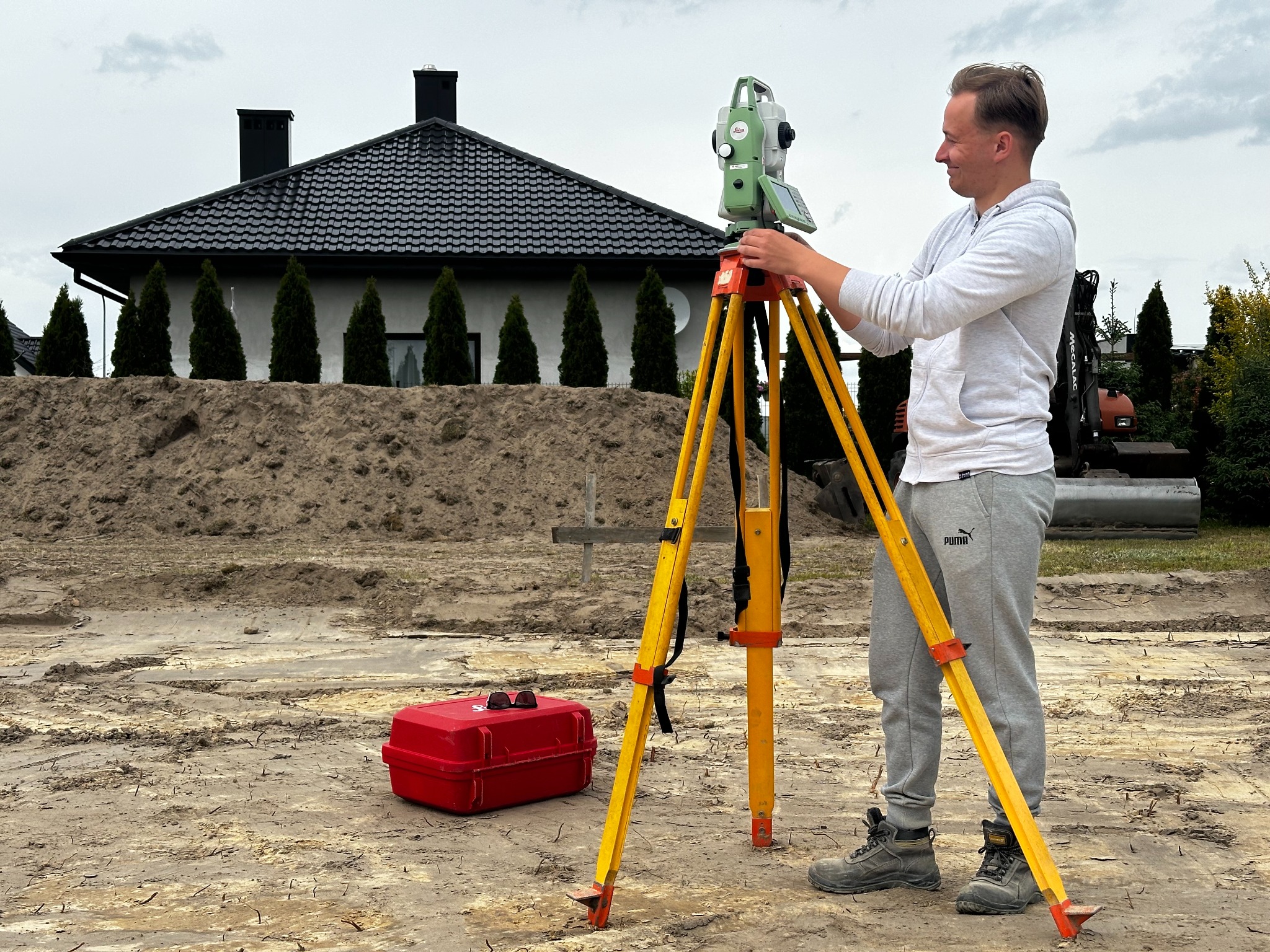 Mężczyzna obsługuje niwelator na żółtym statywie, w tle dom z czarnym dachem i kopiec ziemi. Czerwona skrzynka narzędziowa leży obok. Prace pomiarowe w toku.