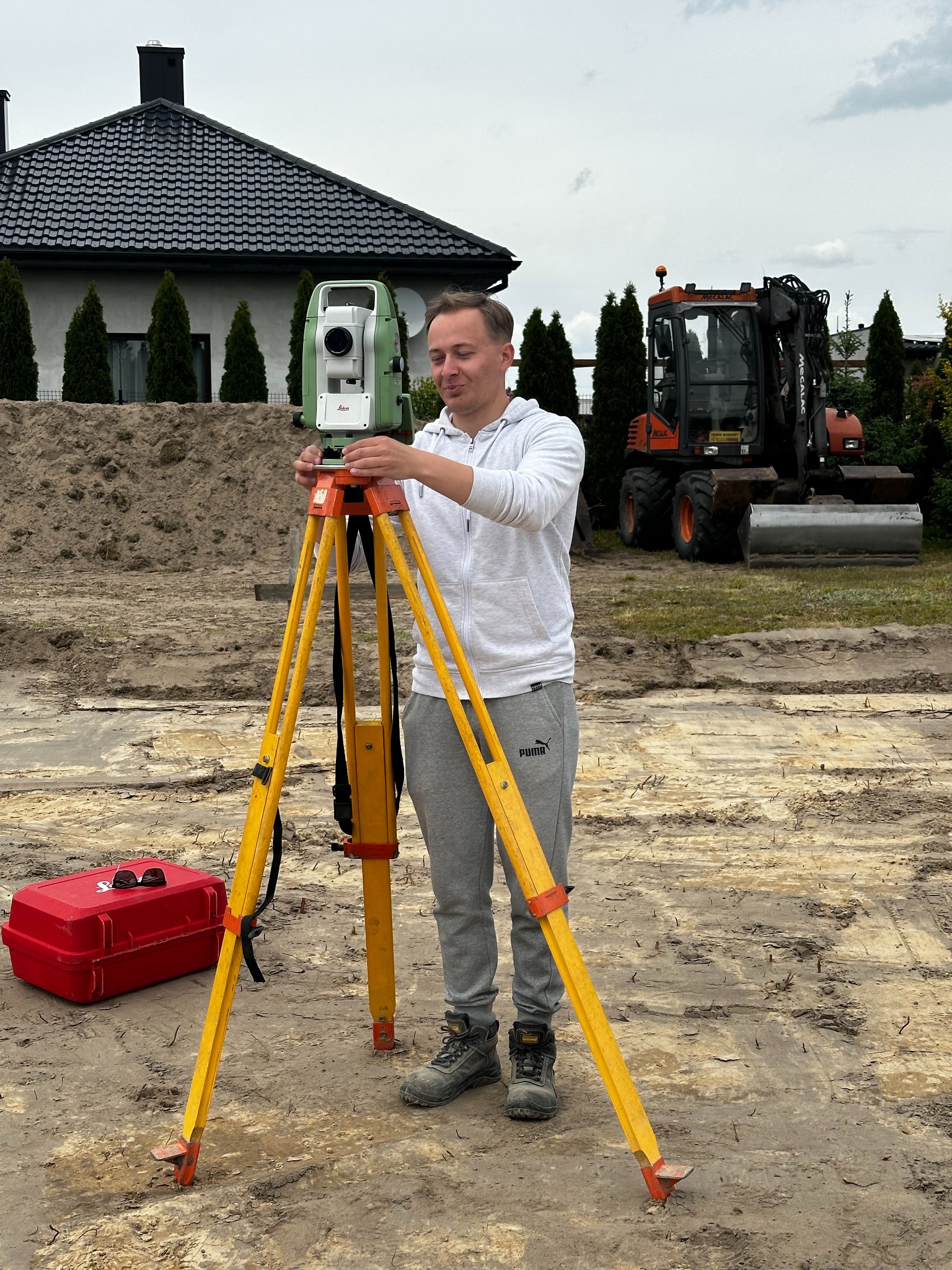 Młody mężczyzna obsługuje niwelator na placu budowy. W tle dom z ciemnym dachem i koparka. Czerwona skrzynka narzędziowa obok statywu.