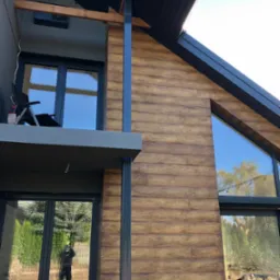 Nowoczesny dom z elewacją imitującą drewno, duże okna z czarnymi ramami, balkon z krzesłem i widokiem na błękitne niebo.