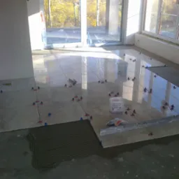 Układanie jasnych płytek podłogowych w słonecznym pomieszczeniu z dużymi oknami, widoczne poziomica, klej i system poziomowania płytek.