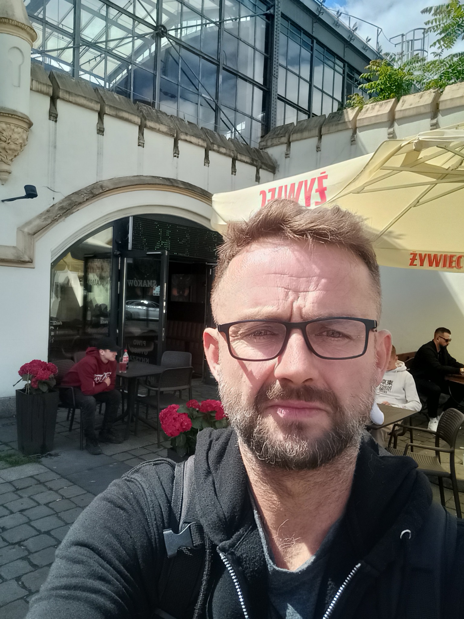 Mężczyzna w okularach na tle kawiarni z przeszklonym dachem i stolikami na zewnątrz. Widoczny fragment parasola i osoby siedzące przy stolikach.