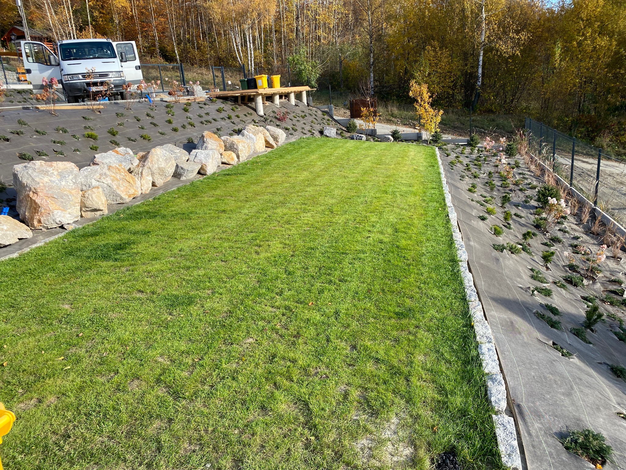 Widok na zielony trawnik z kamiennym obrzeżem, w tle skarpa z agrowłókniną, sadzonkami i drzewami w jesiennej scenerii. Platforma widokowa w tle.
