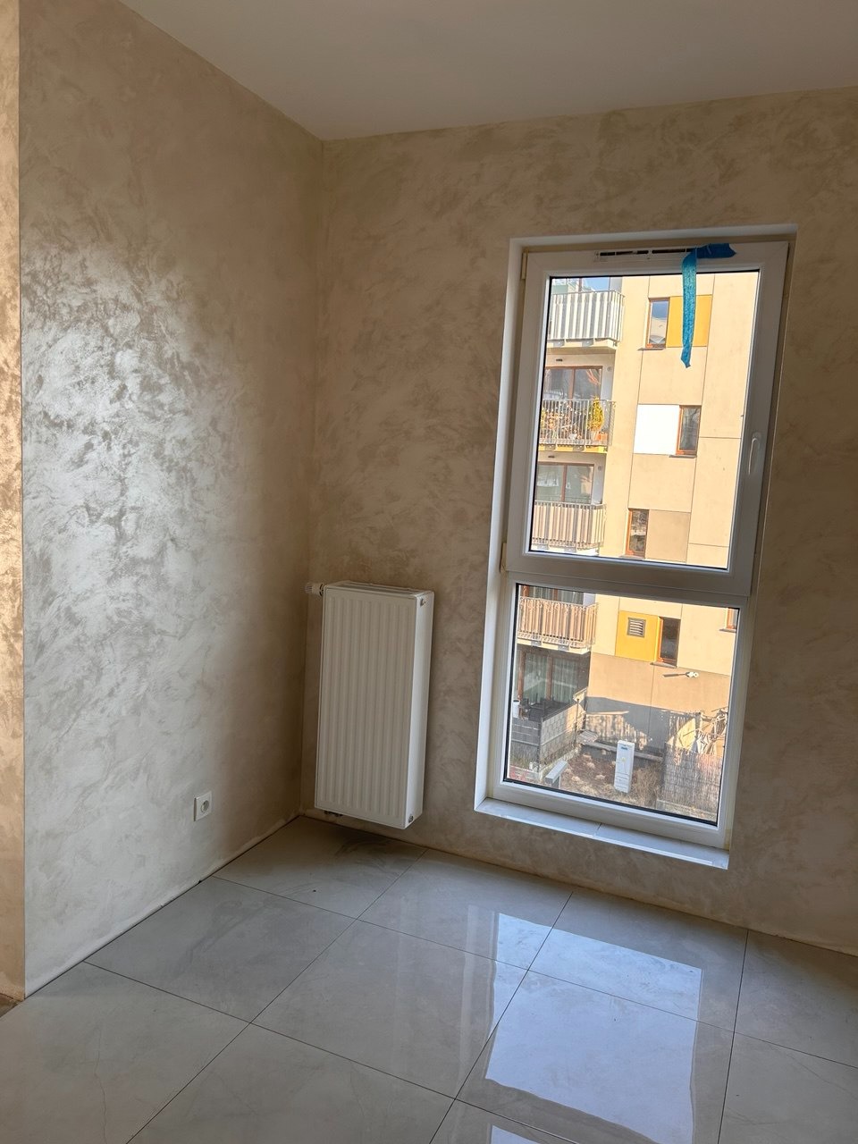 Pokój po wykończeniu ścian z efektem dekoracyjnym, widok na okno z nowym budownictwem. Błyszcząca podłoga z płytek. Grzejnik ścienny.