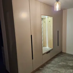 Beżowa szafa na wymiar z siedziskiem i wieszakami, oświetlona od wewnątrz, z długimi, czarnymi uchwytami, na tle marmurowej podłogi i z wiszącą lampą.