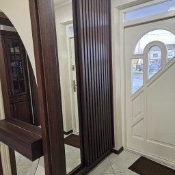 D&A Meble na wymiar Dawid Kosiorkiewicz - Nowoczesna szafa z lustrem i lamelami w przedpokoju, z widocznym fragmentem drzwi wejściowych i podłogi z płytek. Ciemne drewno kontrastuje z jasnymi ścianami.