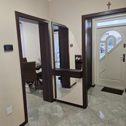 D&A Meble na wymiar Dawid Kosiorkiewicz - Nowoczesny przedpokój z lustrzaną szafą wnękową z ciemnego drewna, odbijającą fragment jadalni. Białe drzwi wejściowe z krzyżem na ścianie. Szare płytki podłogowe.