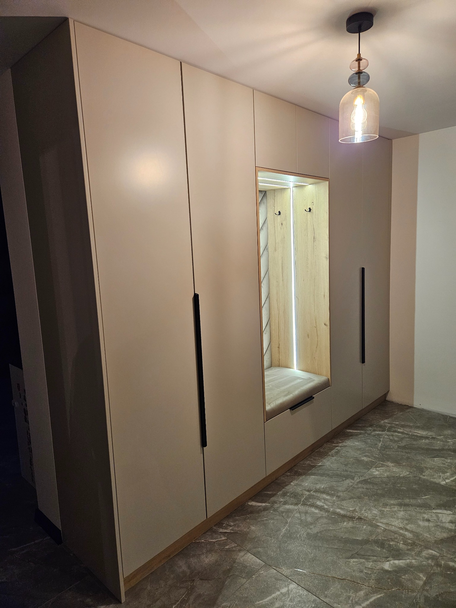 Beżowa szafa na wymiar z siedziskiem i wieszakami, oświetlona od wewnątrz, z długimi, czarnymi uchwytami, na tle marmurowej podłogi i z wiszącą lampą.