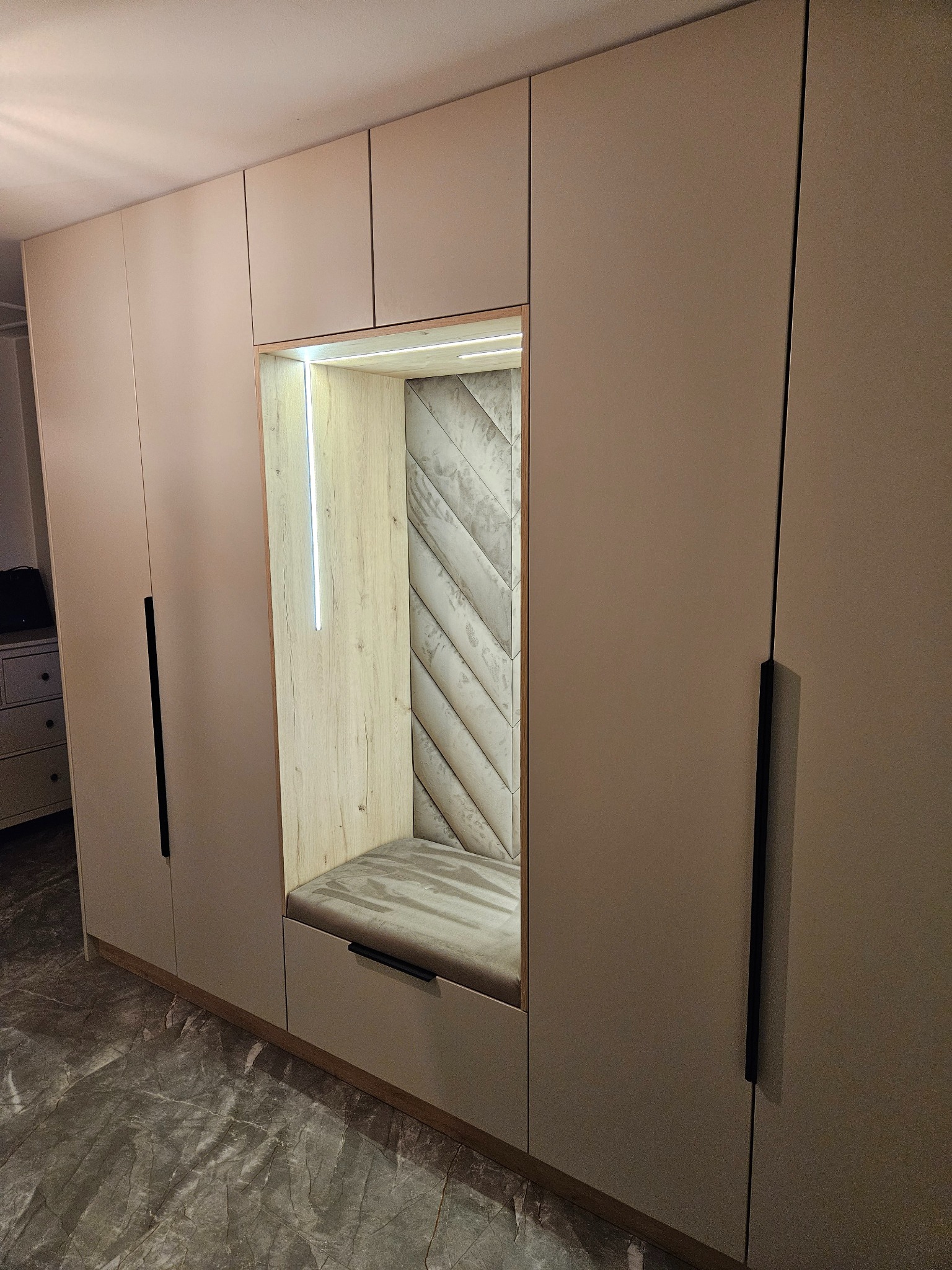 Jasna szafa wnękowa z siedziskiem obitym tkaniną w geometryczny wzór. Wnętrze siedziska oświetlone listwą LED. Czarne, pionowe uchwyty.