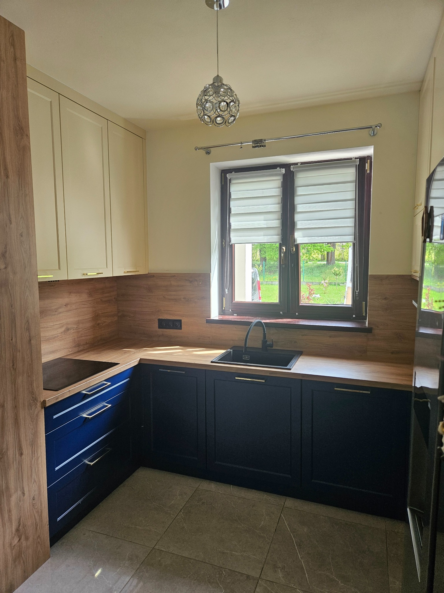 Nowoczesna kuchnia z granatowymi szafkami dolnymi i jasnymi górnymi, drewnianym blatem i backsplashiem. Okno z roletami dzień-noc, widok na zieleń.