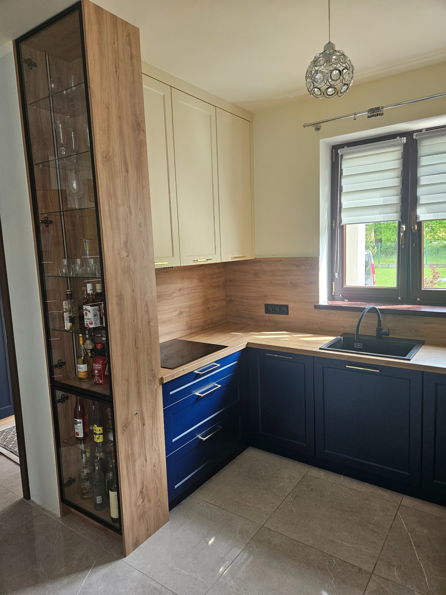 Kuchenna zabudowa: szafki kremowe górne, granatowe dolne, drewnopodobny blat i panel. Wysoka witryna z ekspozycją alkoholi. Nowoczesna lampa sufitowa.