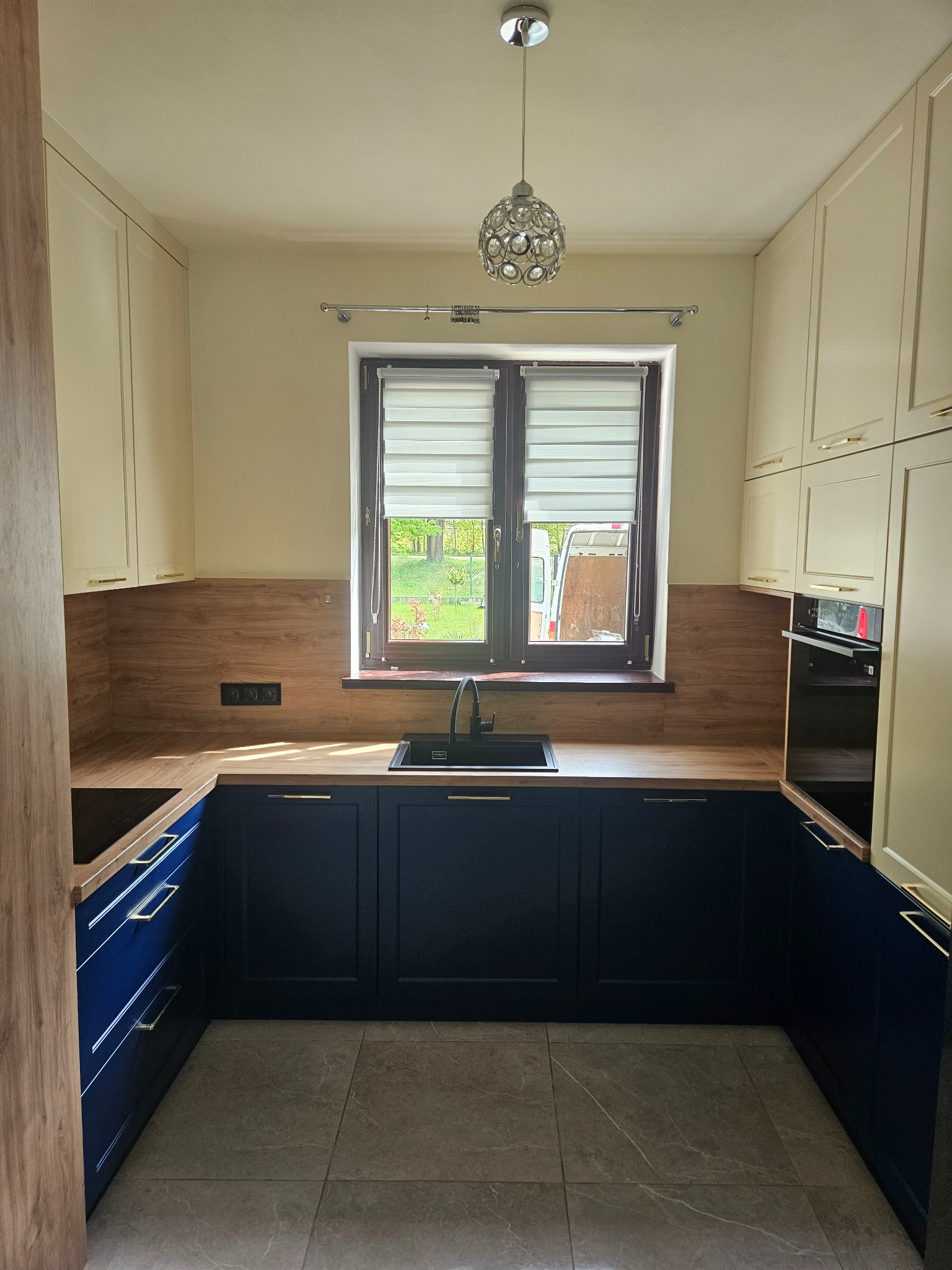 Nowoczesna kuchnia w kształcie litery U z kremowymi szafkami górnymi, granatowymi dolnymi i drewnianym blatem. Widok na okno z roletami i ogród.