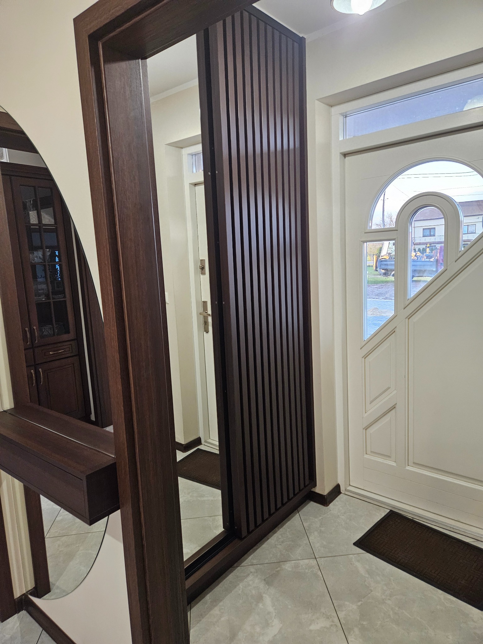 Nowoczesna szafa z lustrem i lamelami w przedpokoju, z widocznym fragmentem drzwi wejściowych i podłogi z płytek. Ciemne drewno kontrastuje z jasnymi ścianami.