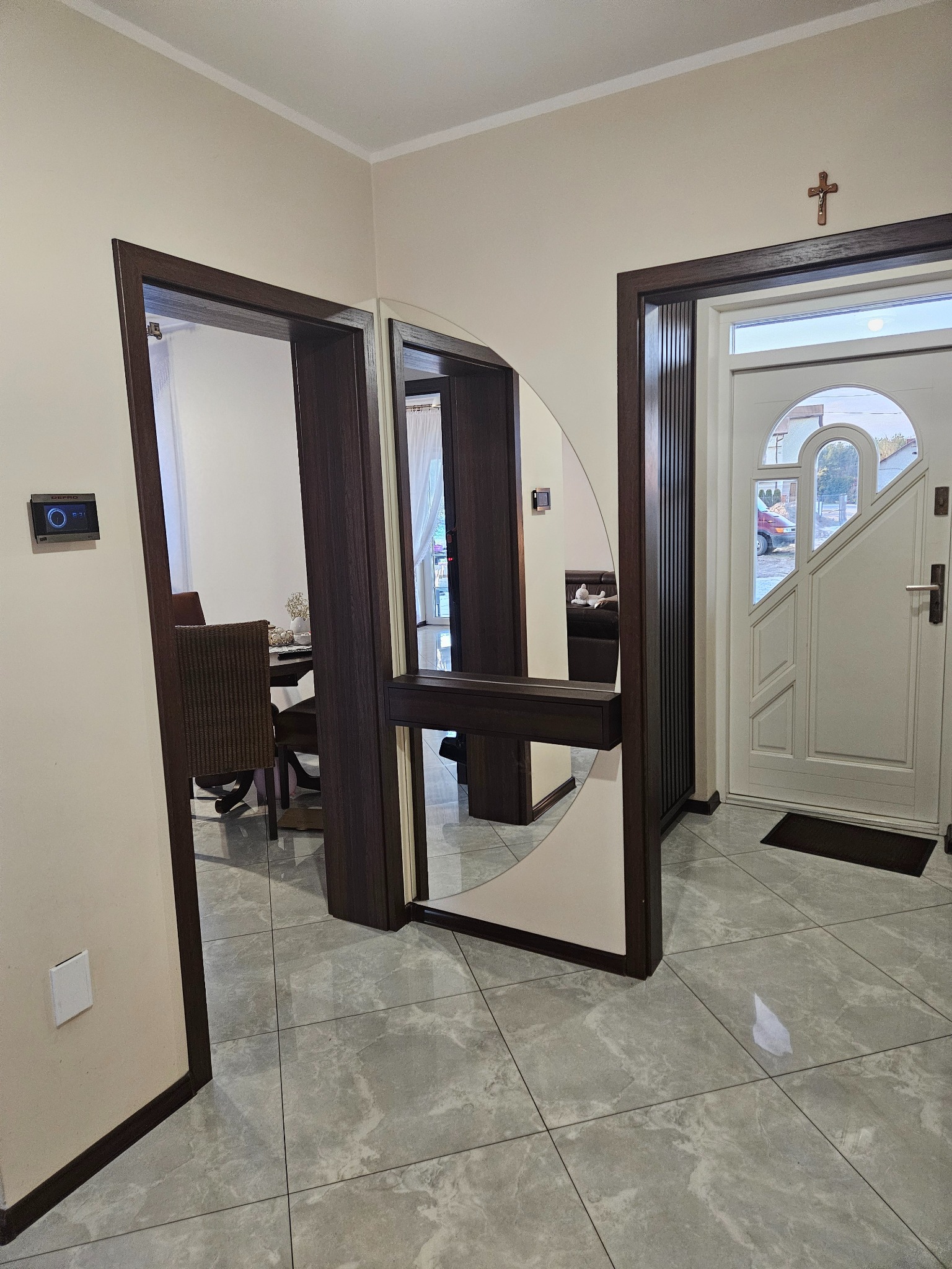 Nowoczesny przedpokój z lustrzaną szafą wnękową z ciemnego drewna, odbijającą fragment jadalni. Białe drzwi wejściowe z krzyżem na ścianie. Szare płytki podłogowe.