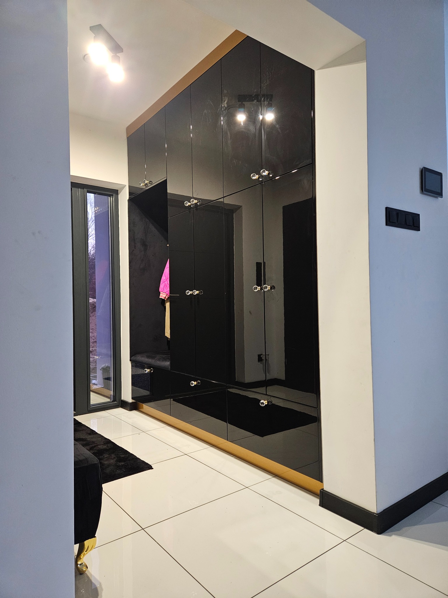 Czarna, lustrzana szafa wnękowa z złotymi akcentami i ozdobnymi uchwytami w przedpokoju. Widoczne odbicie wnętrza szafy i fragment czarnego dywanu.
