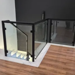 Szklana balustrada z czarną ramą, zabezpieczająca schody z podświetleniem LED. Nowoczesny design z marmurowym wykończeniem podłogi i kontrastującą czarną ścianą.