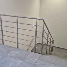 AJ.MOBISPAW - Minimalistyczna balustrada ze stali nierdzewnej na schodach z szarymi płytkami. Ujęcie z góry, widoczne fragmenty klatki schodowej i ściany. Nowoczesny design.