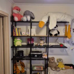 Czarny metalowy regał z drewnianymi półkami, na których stoją książki, zabawki i dekoracje. U góry lampa w kształcie kwiatów.