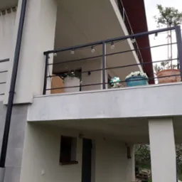 Nowoczesna, czarna balustrada balkonowa z oświetleniem żarówkowym, donice z kwiatami, widok z dołu, jasna elewacja budynku z szarą okładziną.