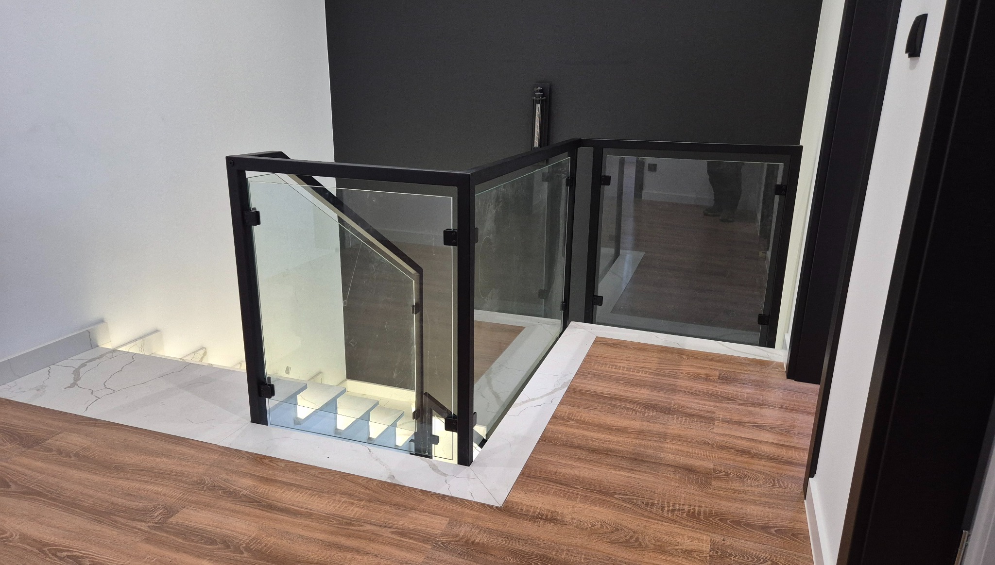 Szklana balustrada z czarną ramą, zabezpieczająca schody z podświetleniem LED. Nowoczesny design z marmurowym wykończeniem podłogi i kontrastującą czarną ścianą.