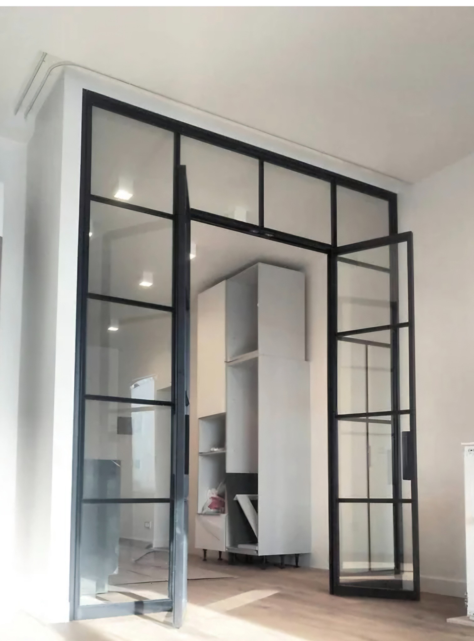 Nowoczesna, przeszklona konstrukcja z czarnymi profilami, tworząca ściankę działową z drzwiami, z widocznym fragmentem zabudowy kuchennej w tle.