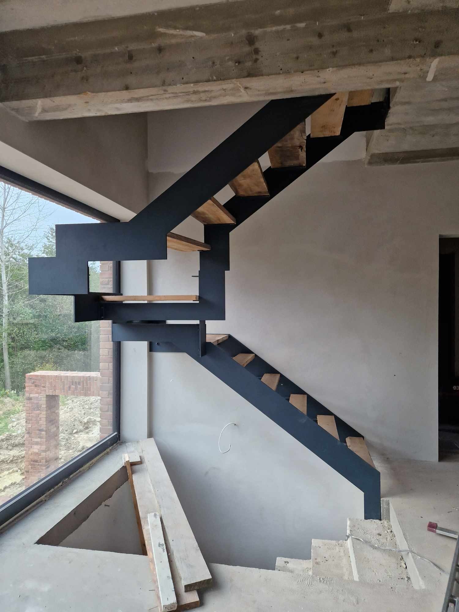 Nowoczesne schody metalowe z drewnianymi stopniami, w surowym wnętrzu budynku w trakcie budowy, z dużym oknem w tle.