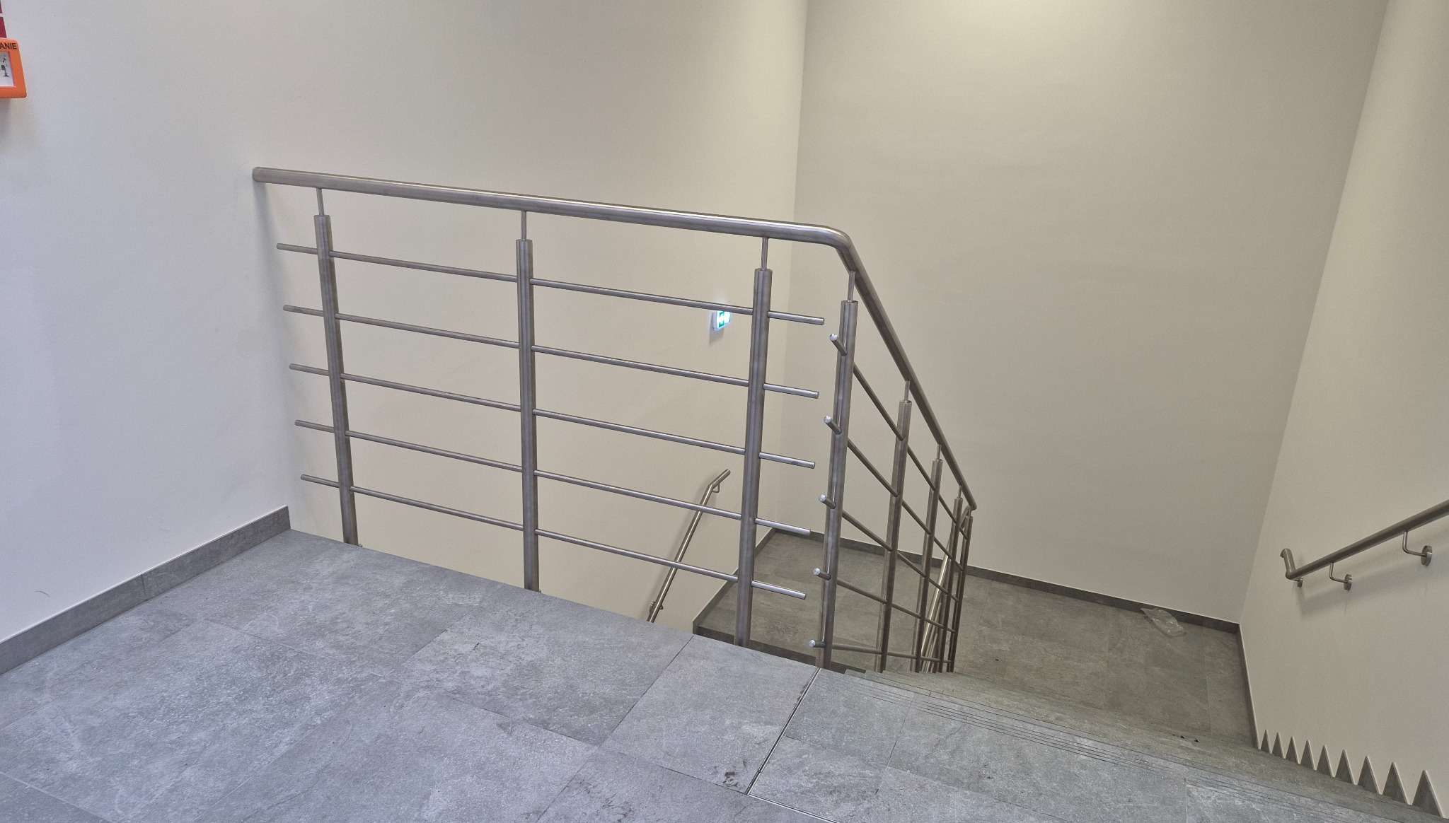 Minimalistyczna balustrada ze stali nierdzewnej na schodach z szarymi płytkami. Ujęcie z góry, widoczne fragmenty klatki schodowej i ściany. Nowoczesny design.