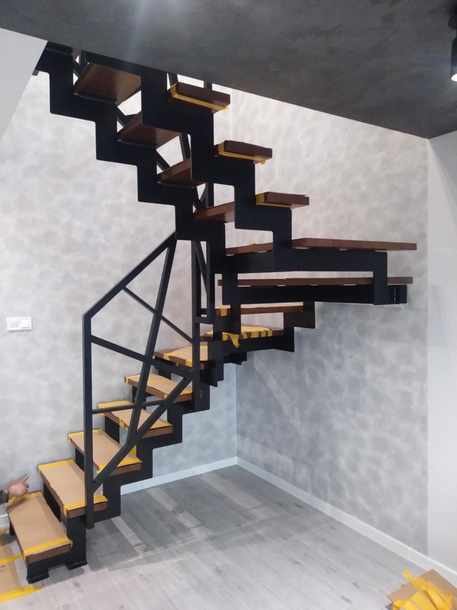 Nowoczesne schody metalowe z drewnianymi stopniami, zabezpieczone taśmą malarską. Minimalistyczny design, szara ściana i podłoga, widok z dołu na konstrukcję.