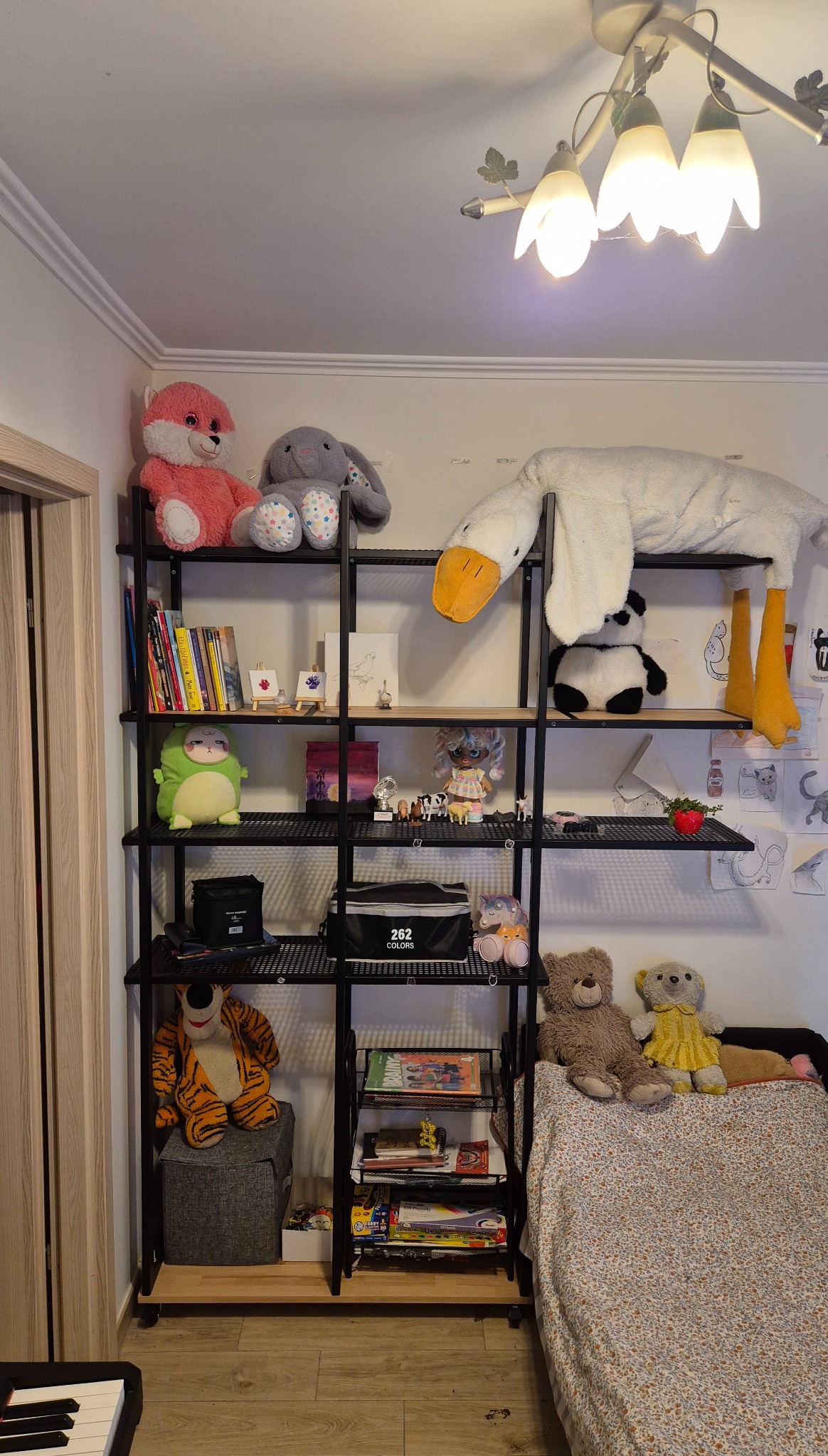 Czarny metalowy regał z drewnianymi półkami, na których stoją książki, zabawki i dekoracje. U góry lampa w kształcie kwiatów.