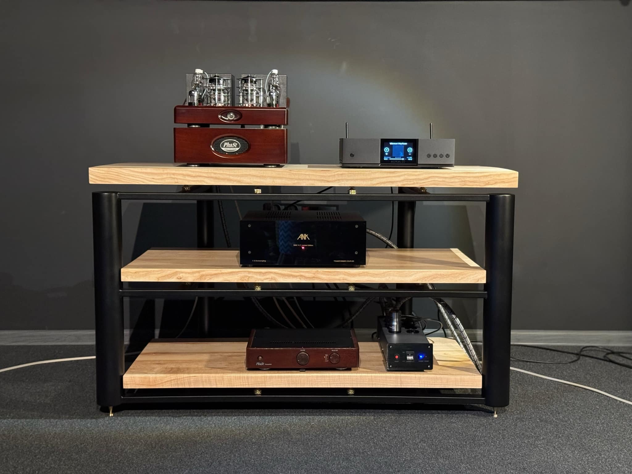 Metalowy regał audio z drewnianymi półkami, na których ustawione są wzmacniacze lampowe i inne komponenty audiofilskie. Czarny stelaż, jasne drewno półek, szary dywan.