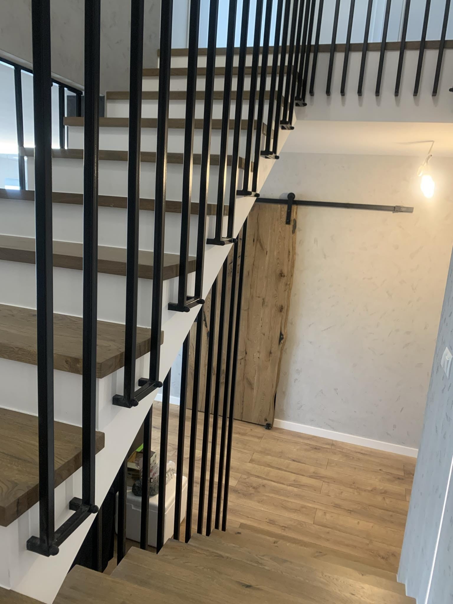 Nowoczesne schody wewnętrzne z drewnianymi stopniami i czarną, metalową balustradą na tle przesuwanych drzwi stodołowych i parkietu. Minimalistyczny design.