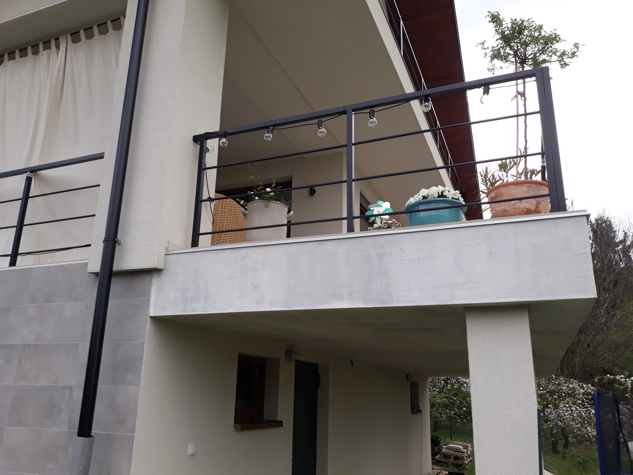 Nowoczesna, czarna balustrada balkonowa z oświetleniem żarówkowym, donice z kwiatami, widok z dołu, jasna elewacja budynku z szarą okładziną.